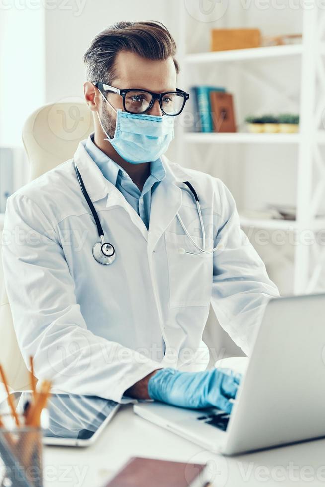 Concentrated young man in white lab coat and protective mask working using laptop while sitting indoors photo