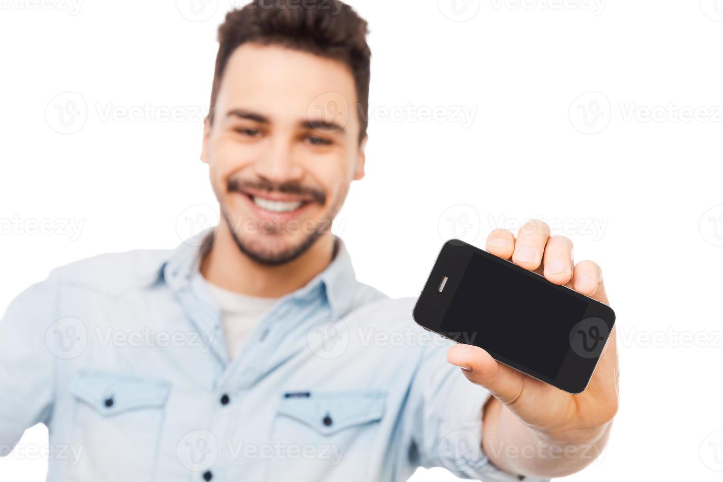 mira mi nuevo aparato joven alegre que muestra el teléfono móvil y sonríe mientras está de pie contra el fondo blanco foto