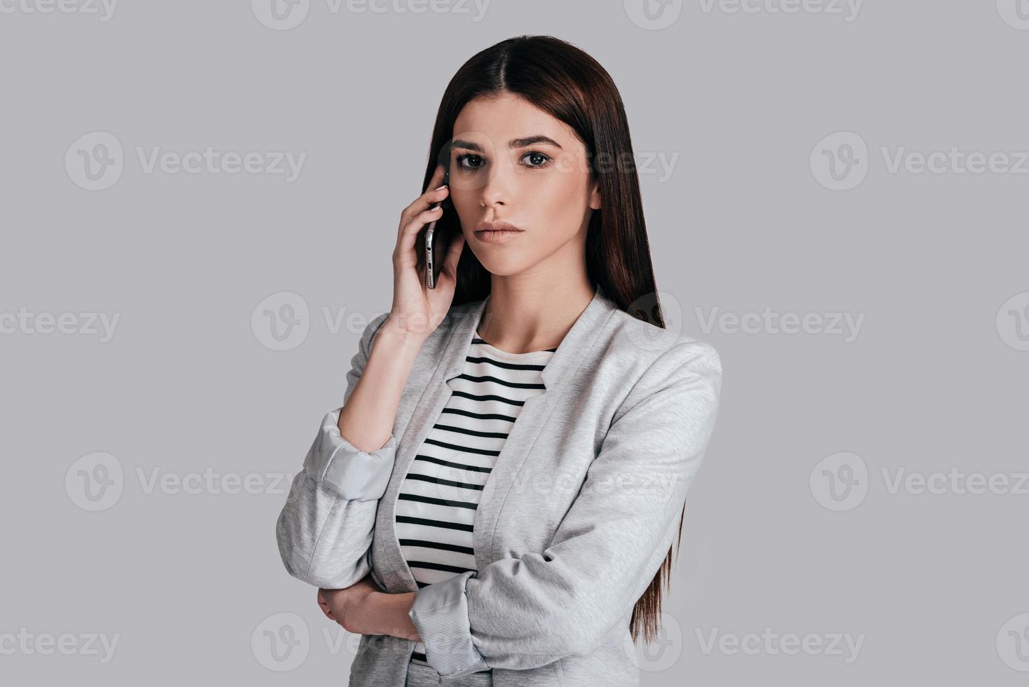 importante charla de negocios. mujer joven hermosa en ropa casual elegante hablando por teléfono y mirando a la cámara con cara seria mientras está pie contra el fondo gris 13581309 Foto