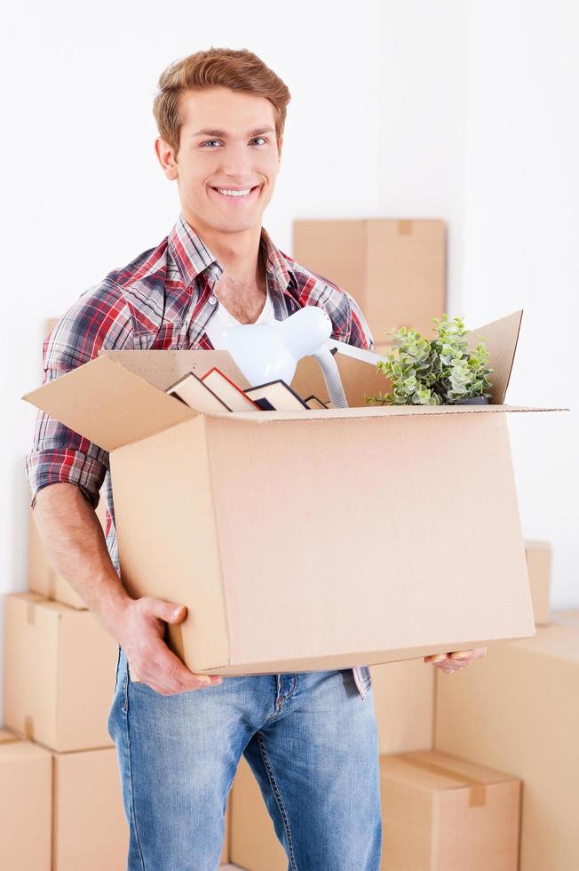 Moving to a new house. Handsome young man holding a cardboard box and smiling at camera while other carton boxes laying on background photo