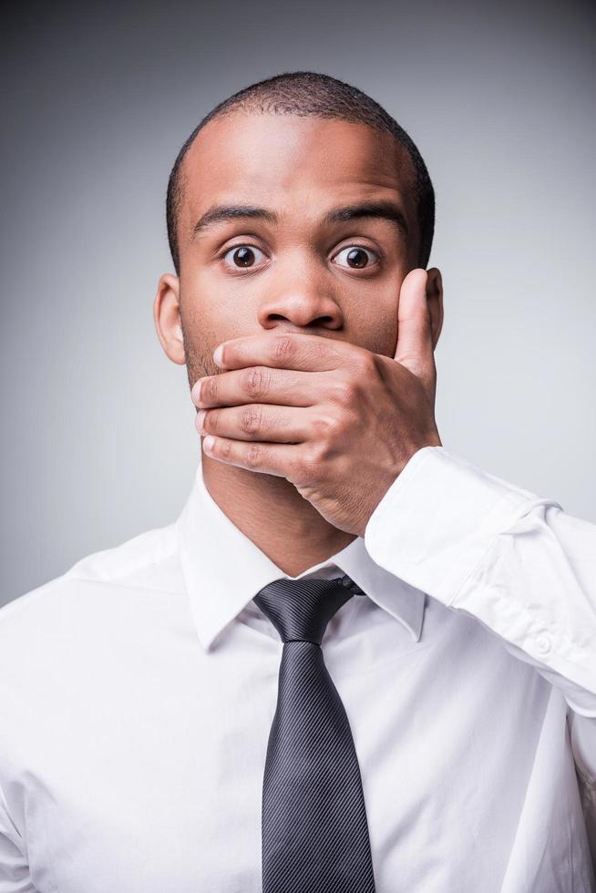 Saying no evil. Shocked young African man in shirt and tie covering mouth with hand while standing against grey background photo