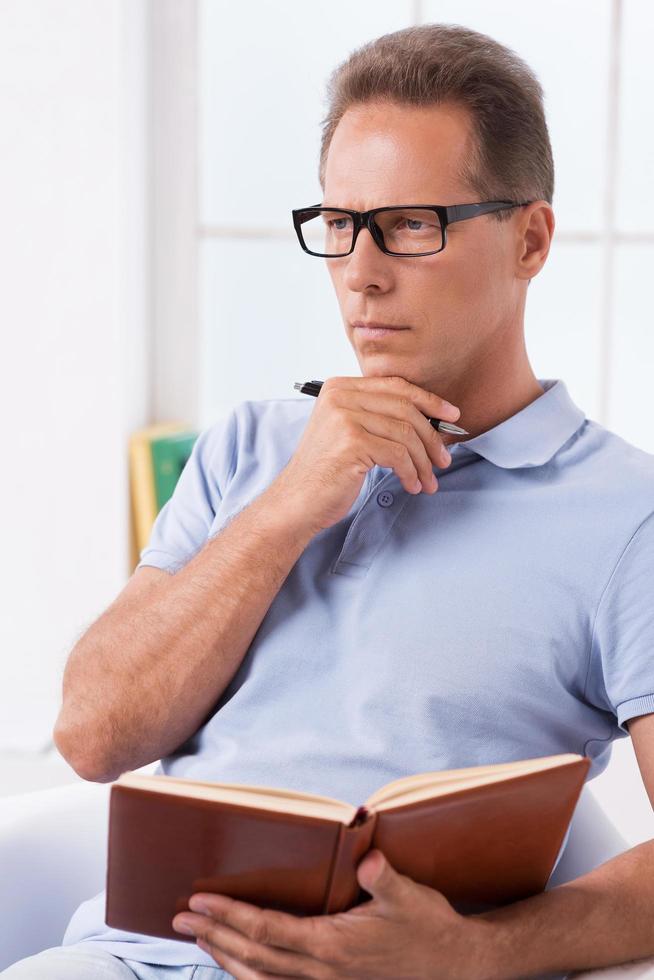 I need some fresh ideas. Thoughtful mature man holding note pad and touching his chin with pen while sitting on the chair photo