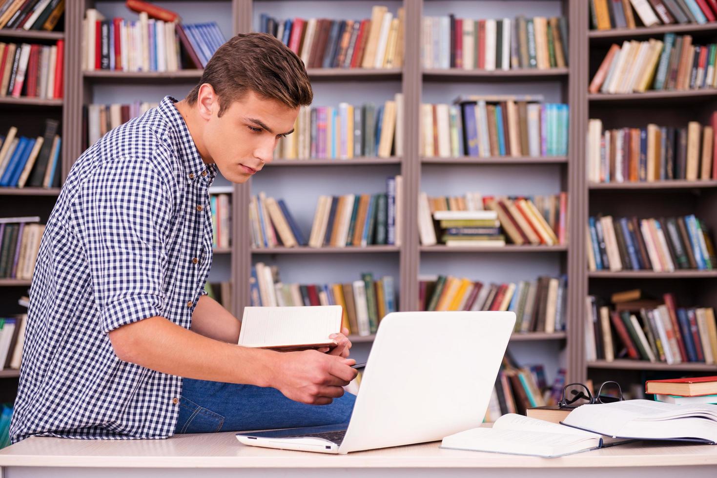 Concentrated on studying. Confident young man holding note pad and looking at laptop while sitting at the desk and in font of bookshelf photo