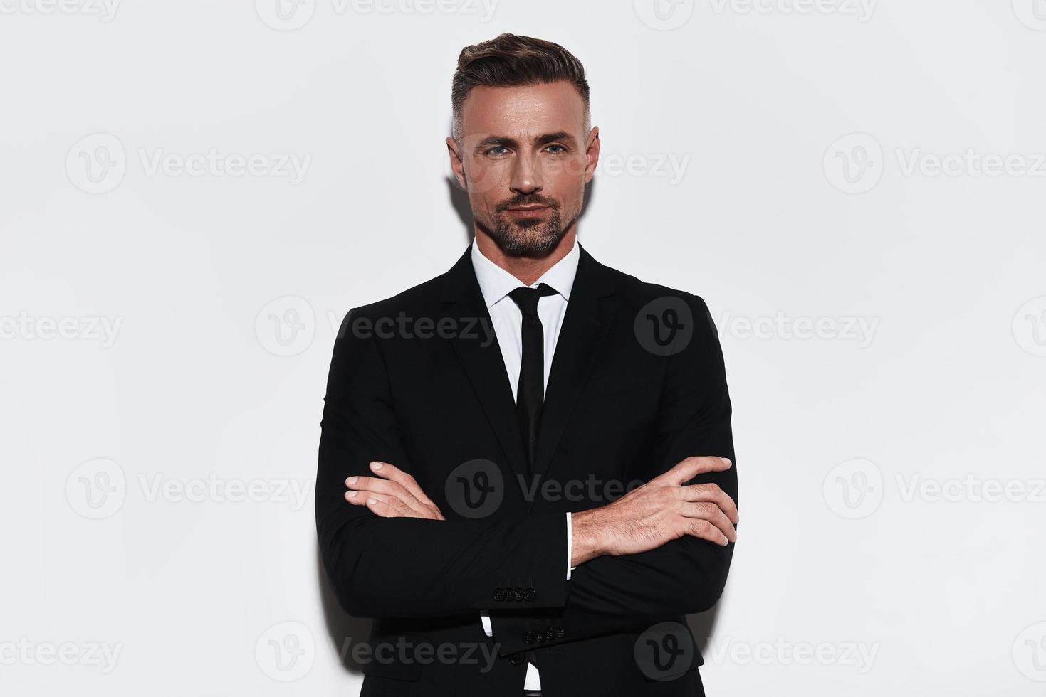 Feeling comfortable in his style. Handsome young man in full suit keeping arms crossed and looking at camera while standing against white background photo