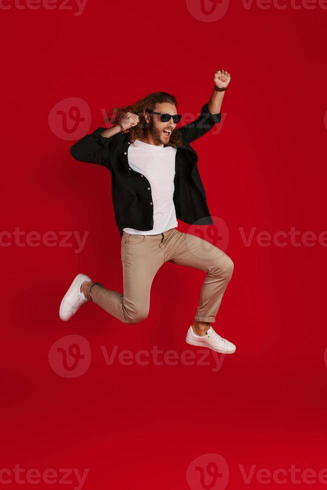 Full length of cheerful young man in casual clothing smiling and gesturing while hovering against red background photo