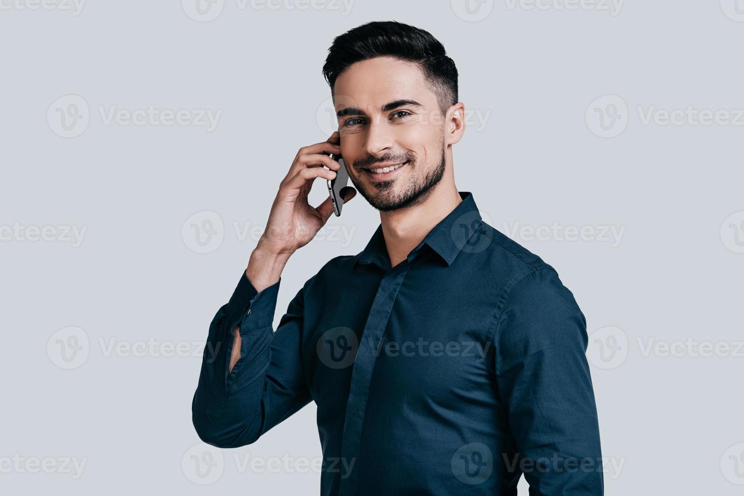 buena charla de negocios. apuesto joven sonriente hablando por teléfono inteligente y mirando a la cámara mientras está de pie contra el fondo gris foto