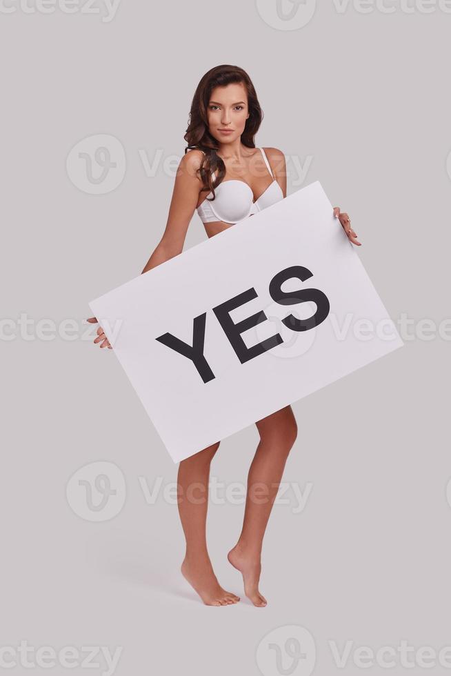Be confident  Full length of attractive young woman covering with a poster and smiling while standing against grey background photo
