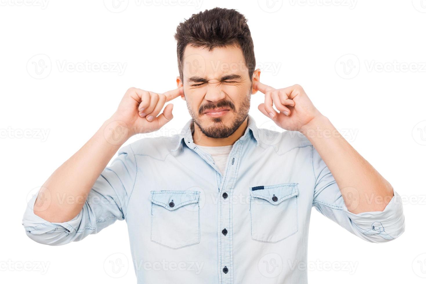 This is too loud Frustrated young man in shirt holding fingers in his ears and keeping eyes closed while standing against white background photo