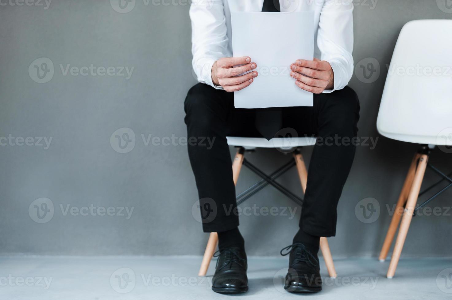 Waiting for interview. Close-up of young man holding paper while sitting on chair against grey background photo