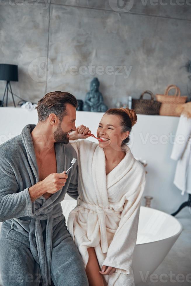 feliz pareja joven en albornoces sonriendo y limpiando los dientes mientras hace la rutina matutina foto