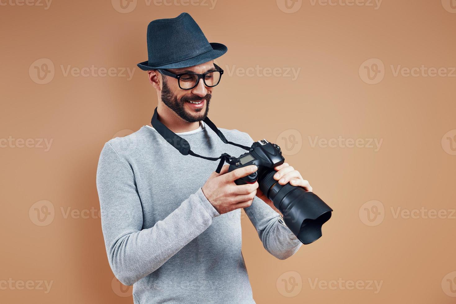 un joven apuesto con ropa informal sonriendo y ajustando la cámara fotográfica mientras se enfrenta a un fondo marrón foto