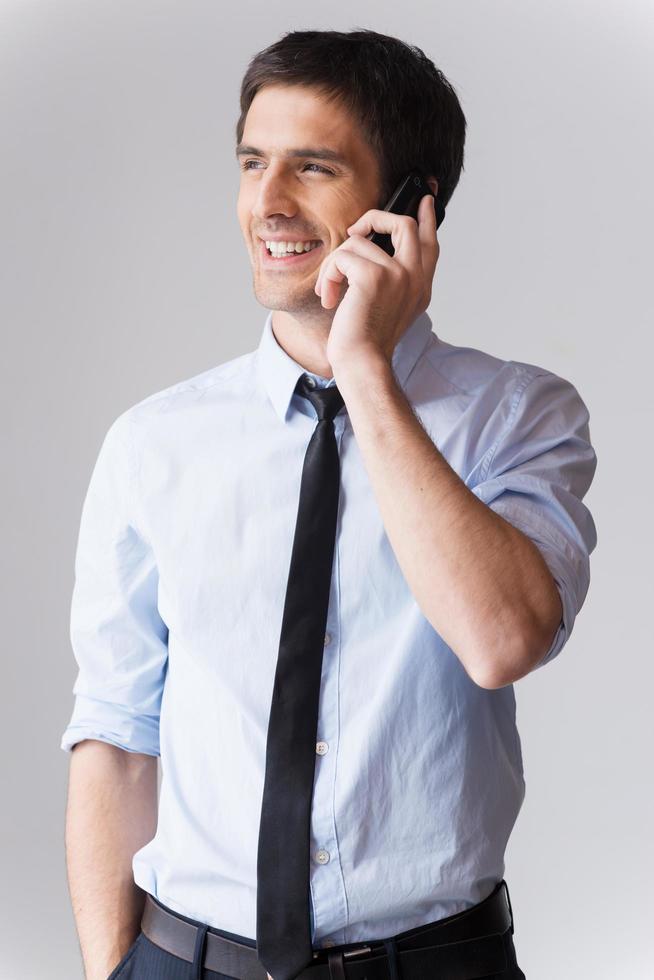 Business talk. Cheerful young man in shirt and tie talking on the mobile phone and smiling while standing against grey background photo