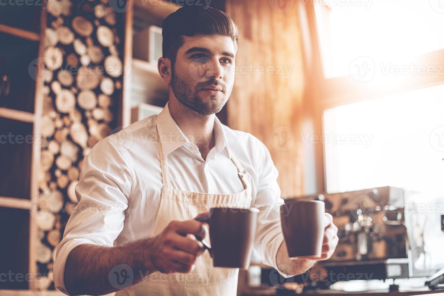 Here you go Young handsome man in apron holding two coffee cups and looking away while standing at cafe photo