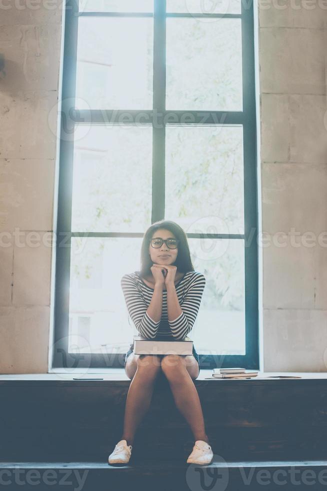 estudiante confiado. hermosa joven mujer de raza mixta que lleva un libro de rodillas y mira la cámara mientras está sentada en el interior y cerca de la ventana grande foto