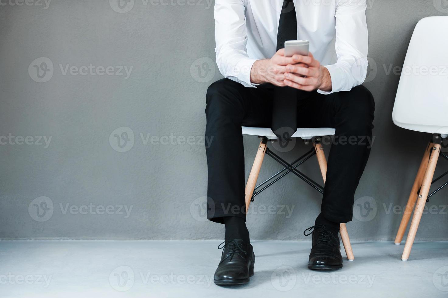 Sending business message. Close-up of young man in shirt and tie holding mobile phone while sitting on chair against grey background photo