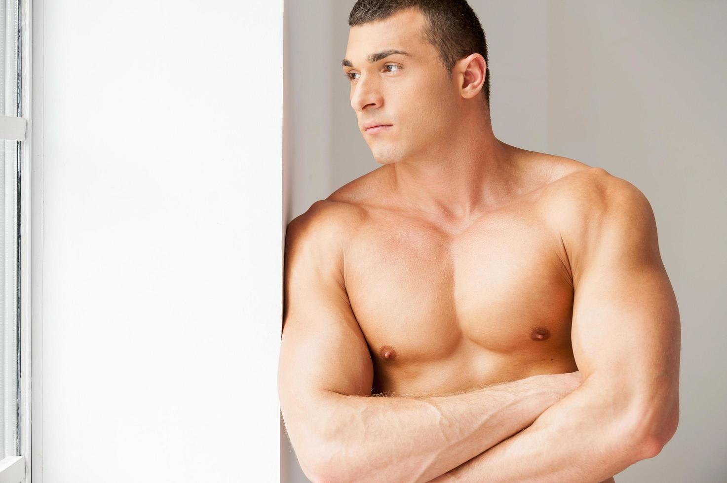 Lost in thoughts. Thoughtful young muscular man looking away while standing near the window photo