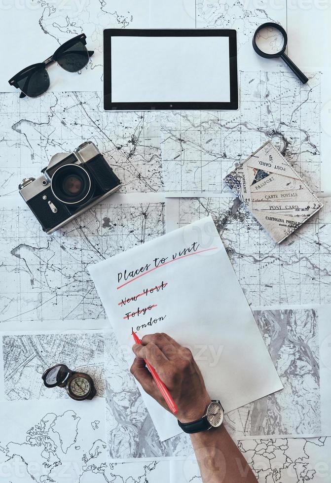 Checklist. Close up top view of man writing down checklist with sunglasses, photo camera, compass, magnifying glass and passport lying on map around