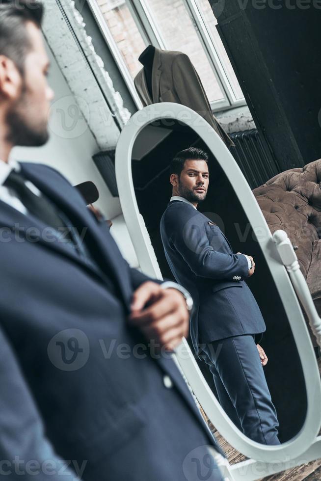 Suit fits perfect. Reflection of handsome young man in full suit adjusting his jacket while standing in front of the mirror indoors photo