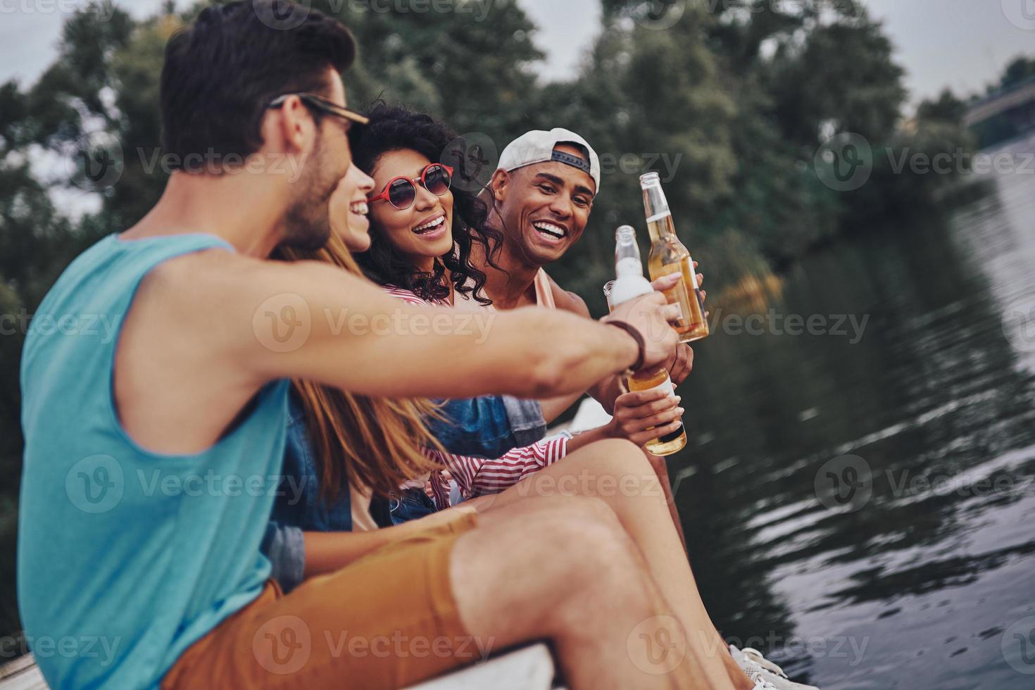 Enjoying time together. Group of happy young people in casual wear smiling and drinking beer while sitting on the pier photo