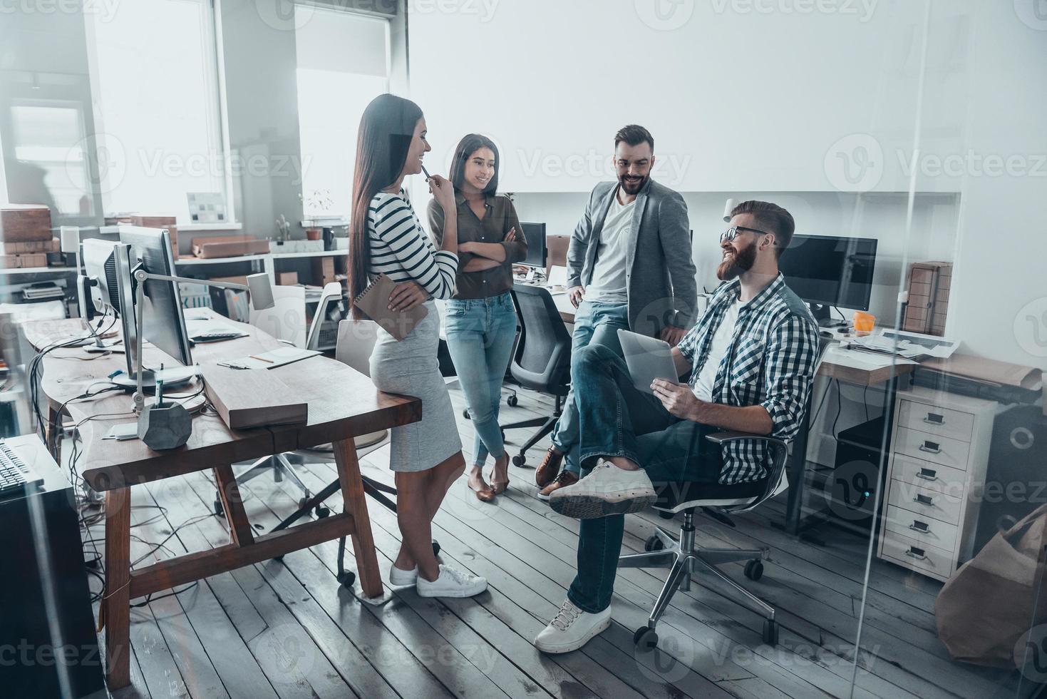 Day meeting. Handsome young man in casual wear and eyeglasses sitting on chair and holding digital tablet while his colleagues standing around him in office photo