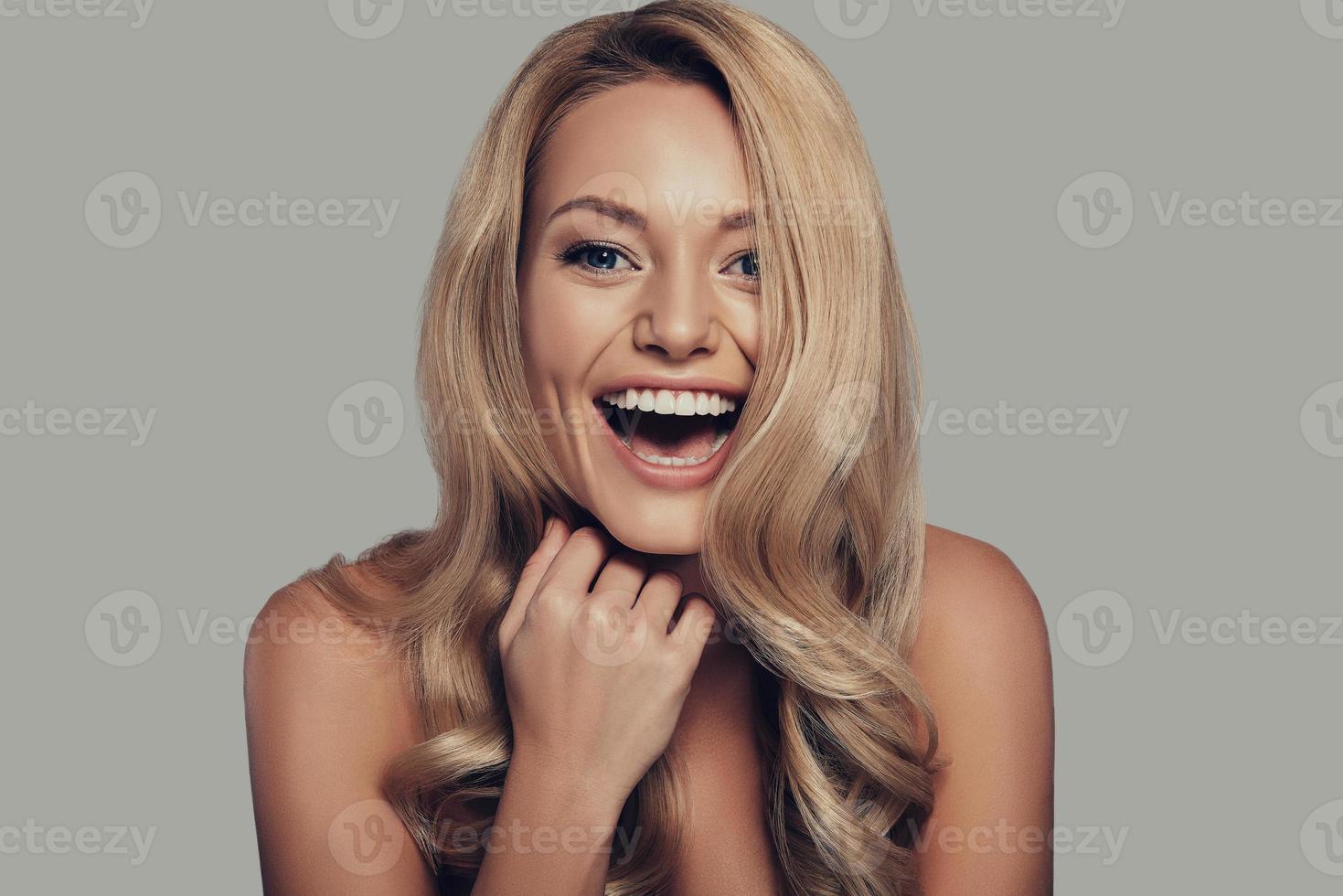 Just happy. Beautiful young woman keeping mouth open and looking at camera while standing against grey background photo