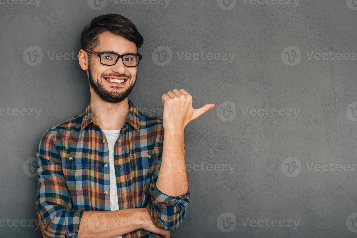 You should go there Cheerful young man in glasses pointing at copy space and looking at camera with smile while standing against grey background photo