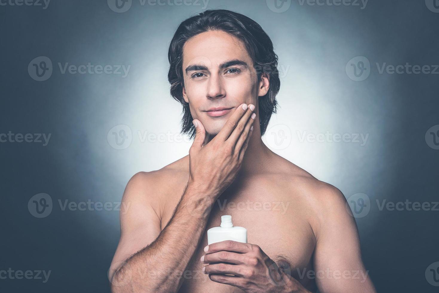 Feeling fresh after shaving. Portrait of handsome young shirtless man looking at camera and applying aftershave lotion on face while standing against grey background photo