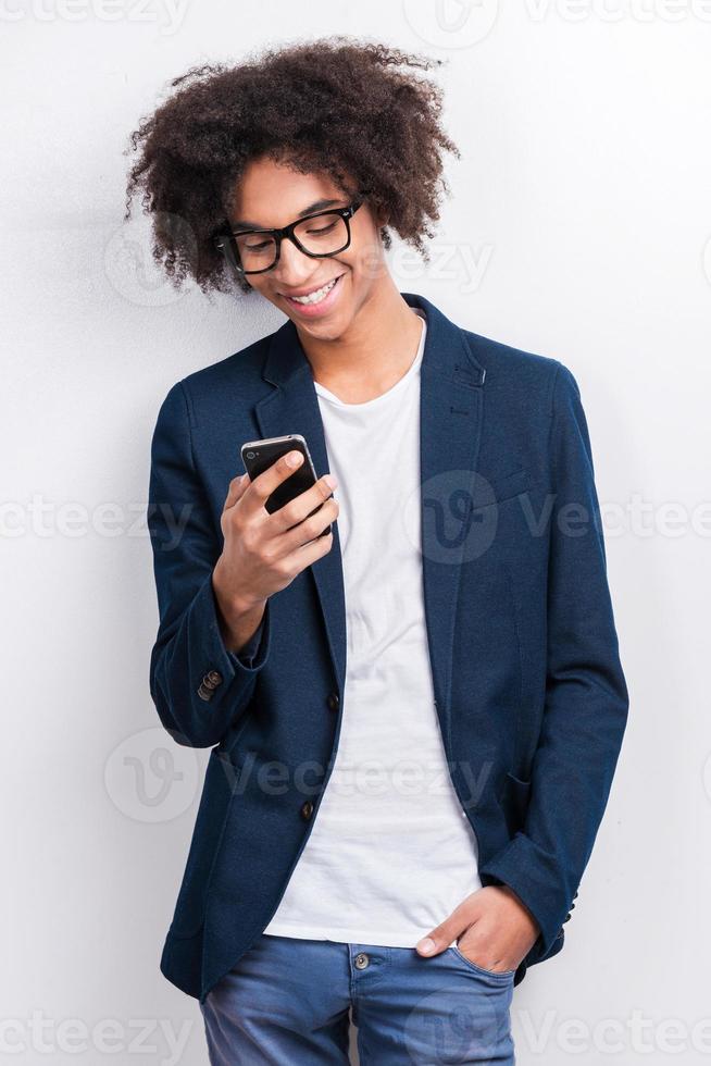 Typing message to friend. Handsome young African man holding mobile phone and smiling while standing against grey background photo