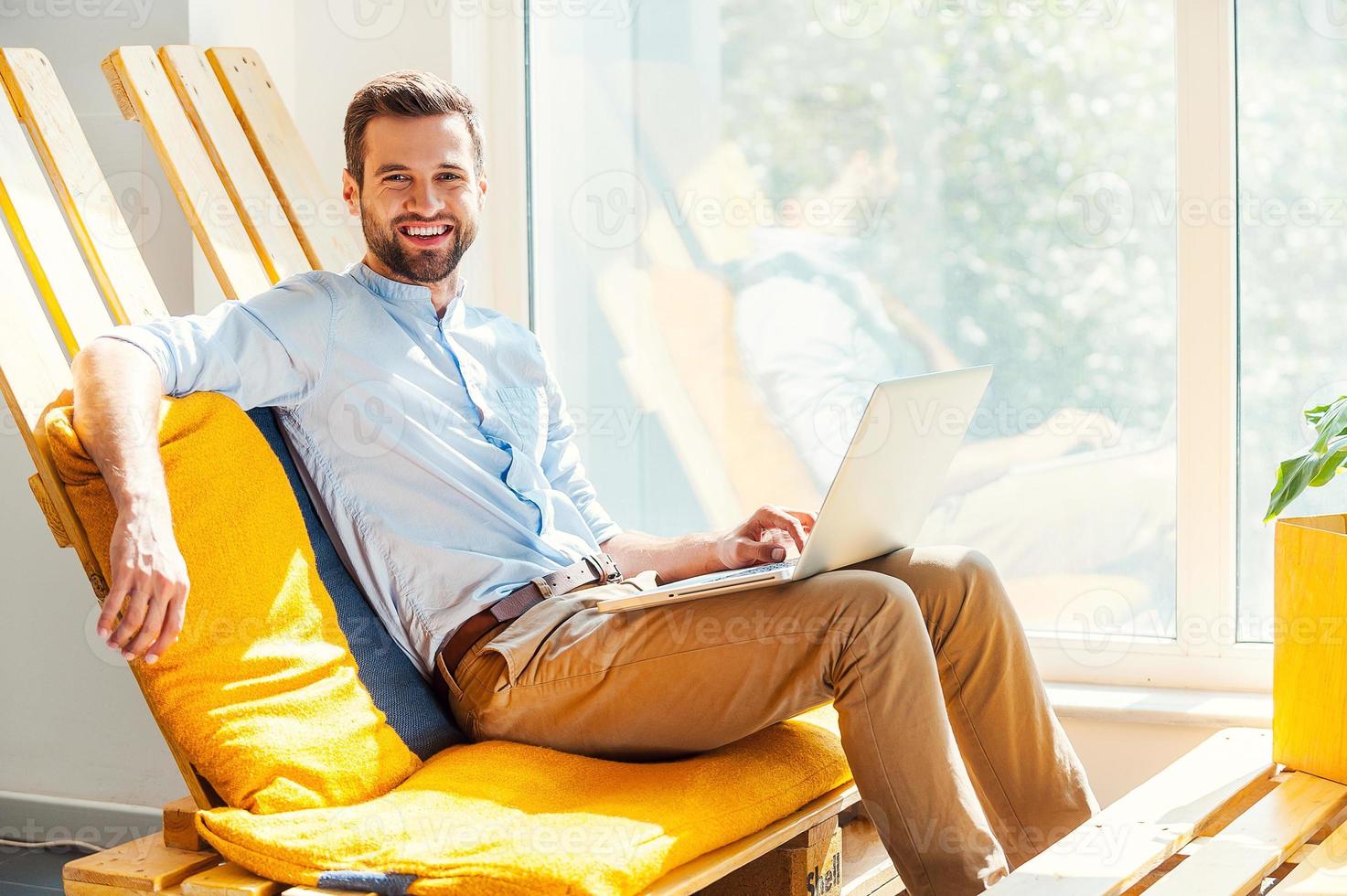 Ready to do business. Happy young man working on laptop and looking at camera while sitting inthe rest area of the office photo
