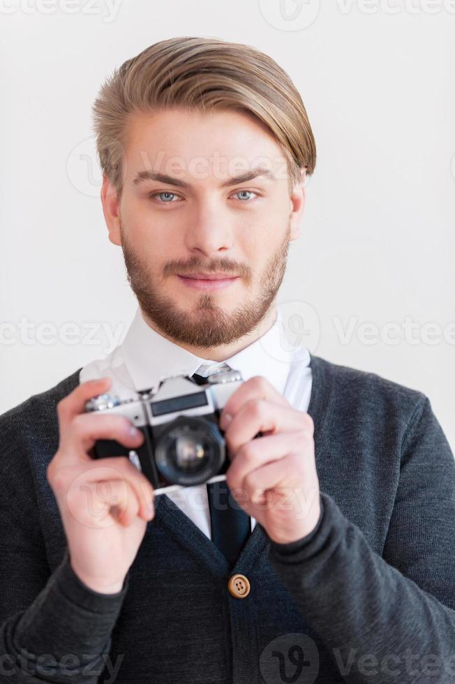 fotógrafo a la antigua. un joven guapo sosteniendo una cámara retro y sonriendo mientras se enfrenta a un fondo gris foto