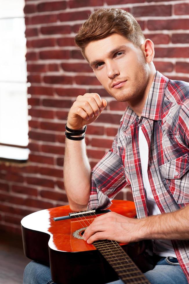 Young and creative. Side view of handsome young man holding acoustic guitar and looking at camera while sitting against brick wall photo