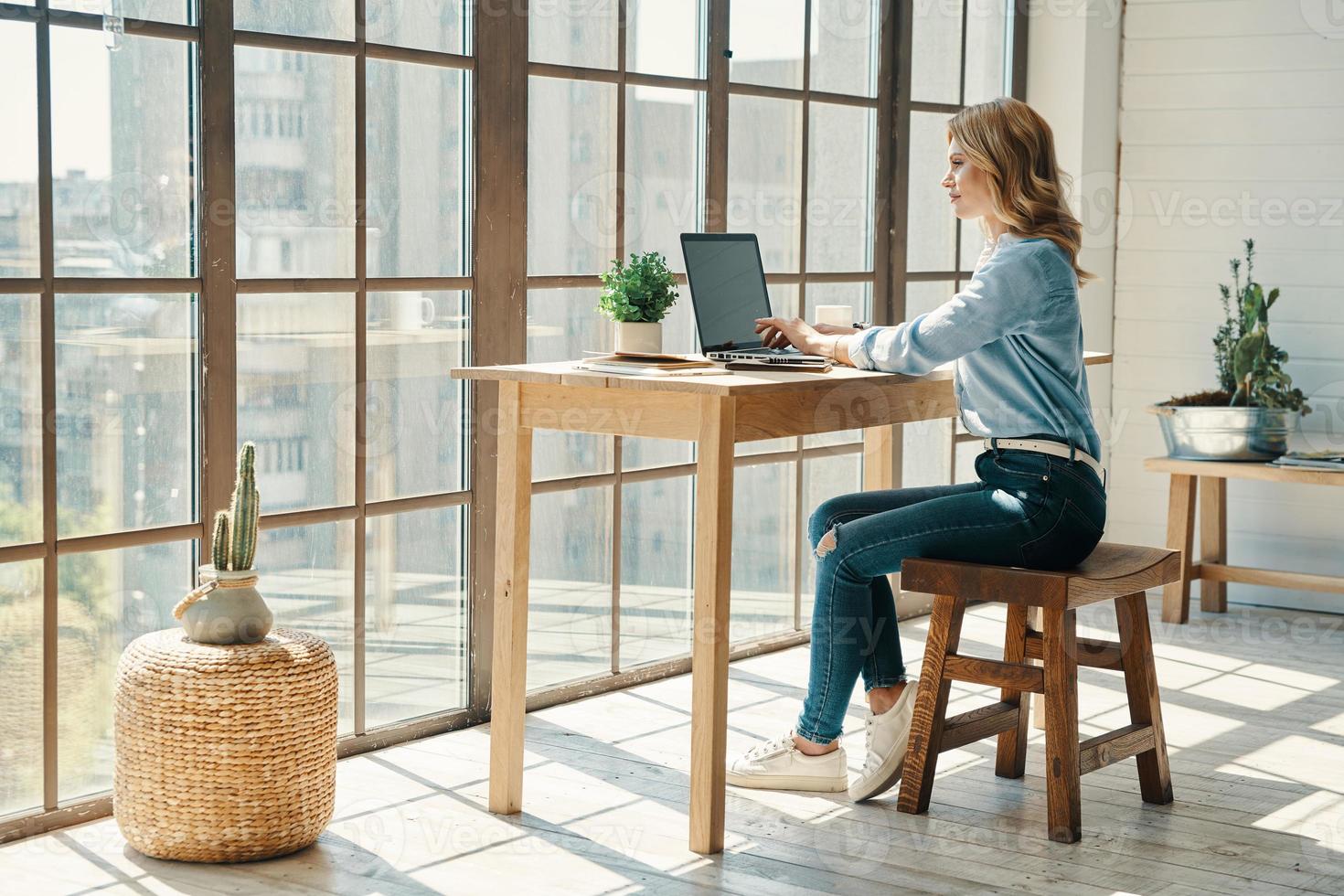 Confident young woman in smart casual wear using laptop while sitting in front of the window photo