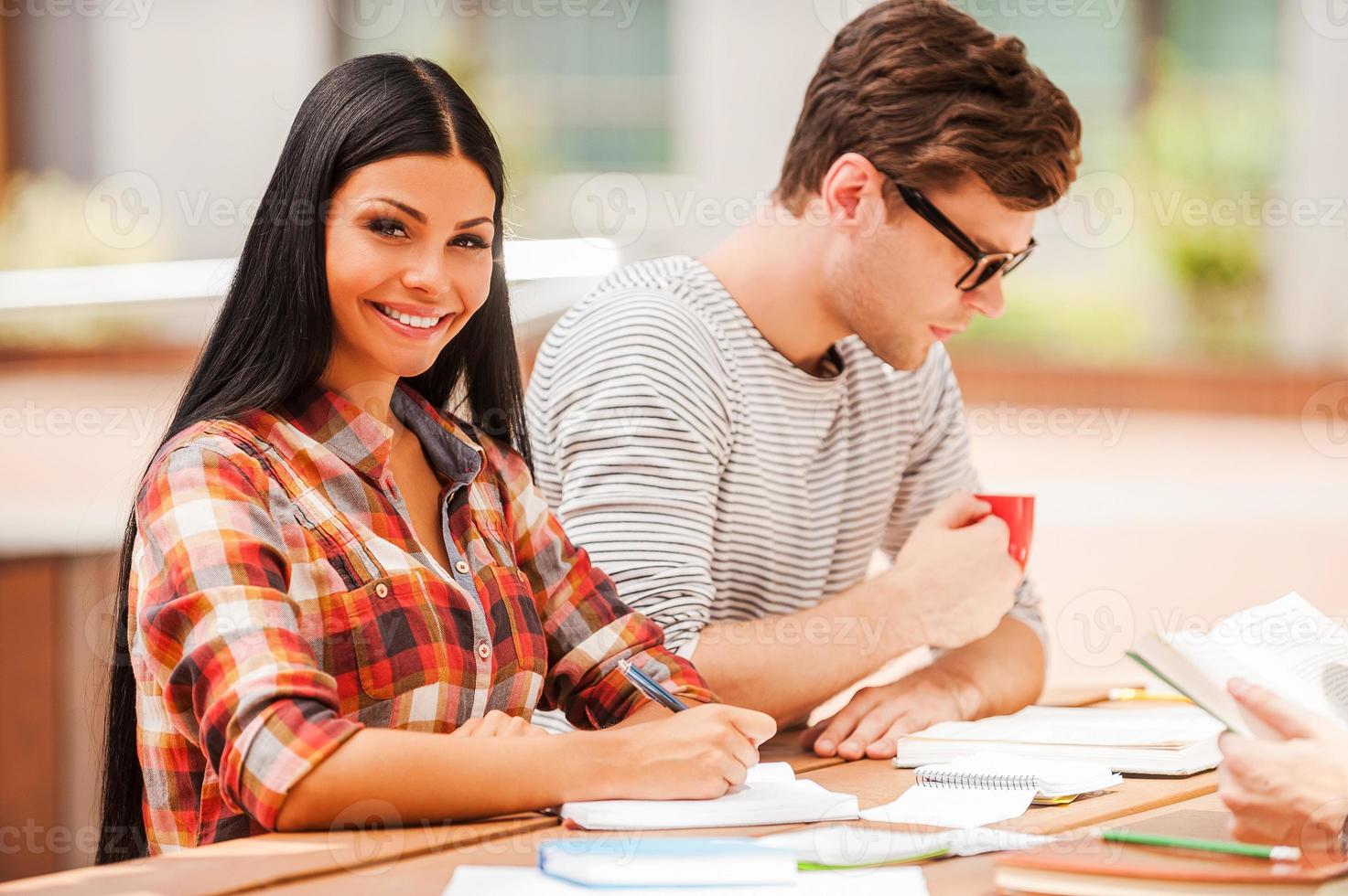 Getting ready for the next class. Smiling young woman writing in note pad and looking at camera while sitting with her friends at the wooden desk outdoors photo