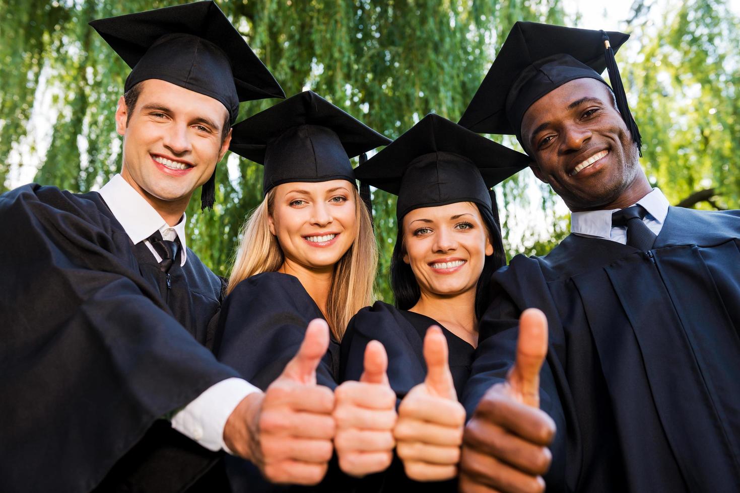Successful graduates. Low angle view of four college graduates in graduation gowns standing close to each other and showing their thumbs up photo