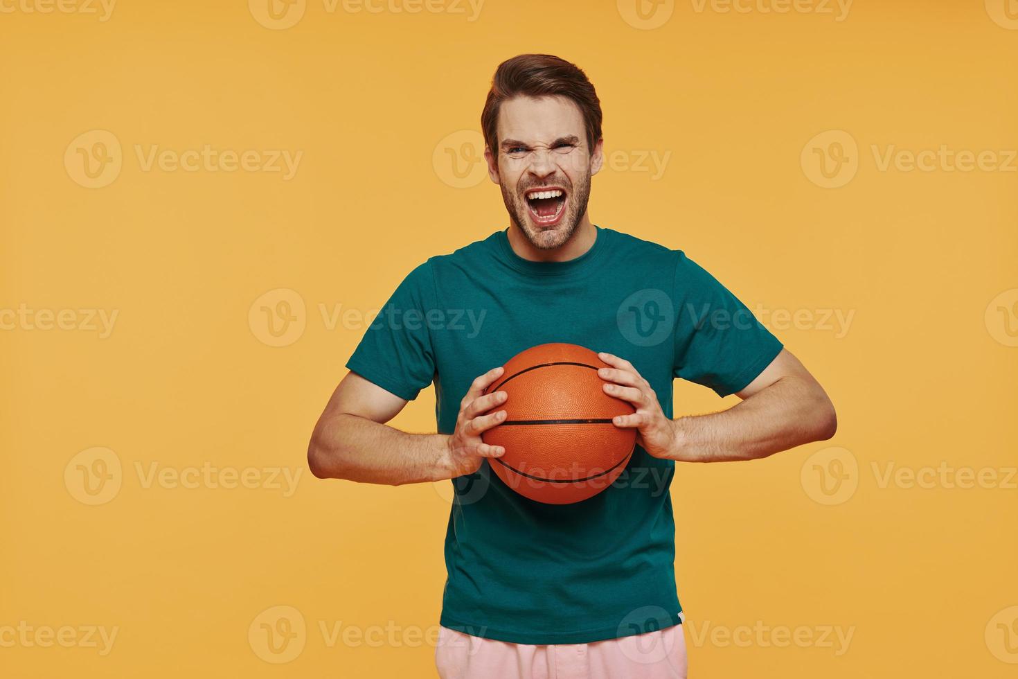 Furious young man holding basketball ball and looking at camera while standing against yellow background photo