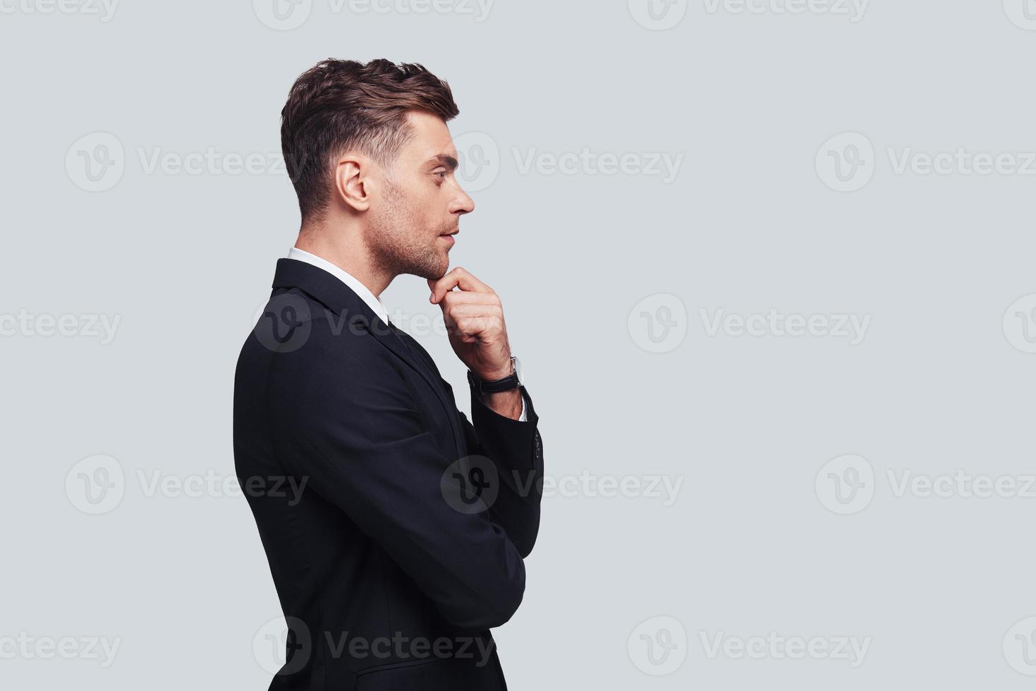 Taking time to think. Serious young man in full suit keeping hand on chin and looking away while standing against grey background photo