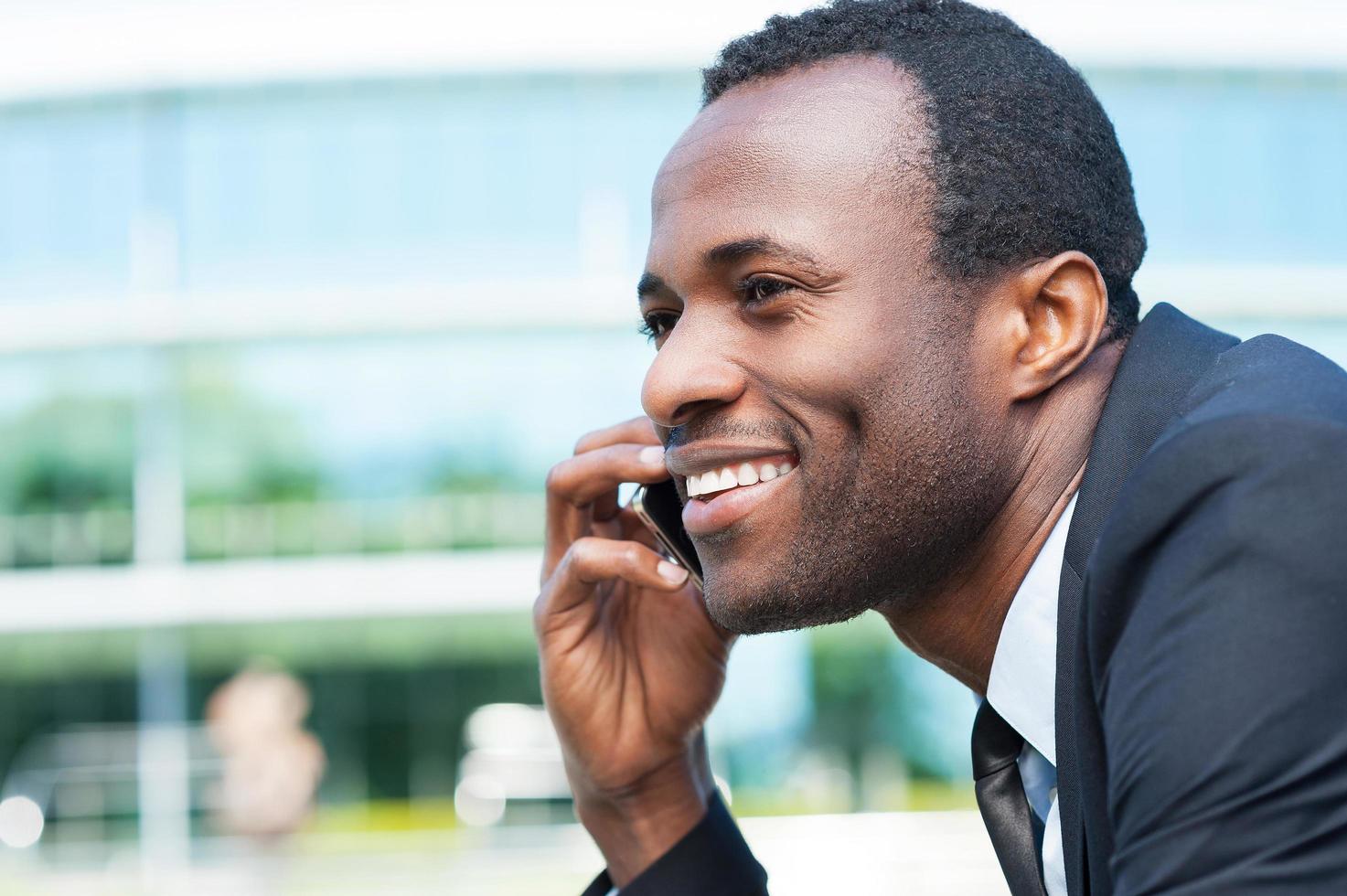 hombre de negocios por teléfono. vista lateral de un apuesto joven africano con ropa formal hablando por teléfono móvil y sonriendo mientras está de pie al aire libre foto