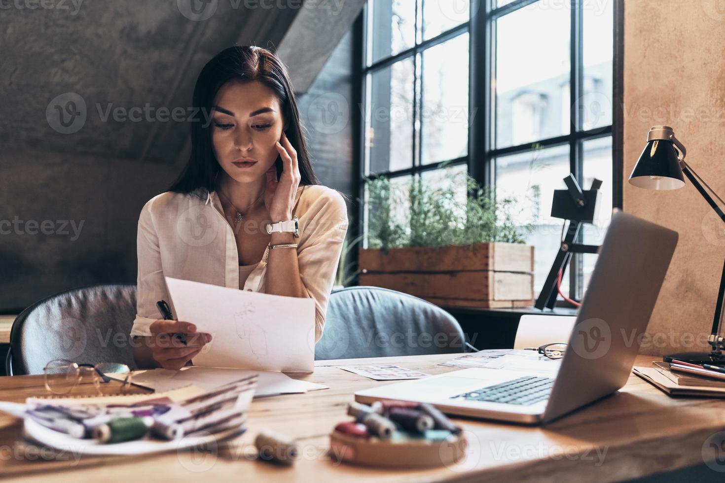 Working on new design. Beautiful young woman holding a paper and looking at it while sitting at the desk in her workshop photo