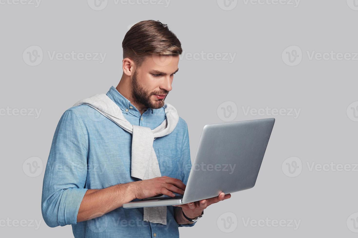 Concentrated at work.  Good looking young man working on his laptop while standing against grey background photo