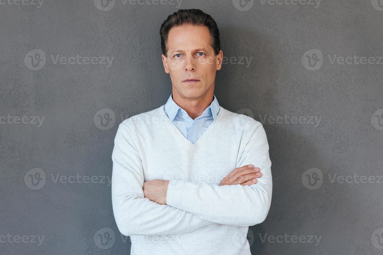 Confident mature man. Confident mature man keeping arms crossed and looking at camera while standing against grey background photo