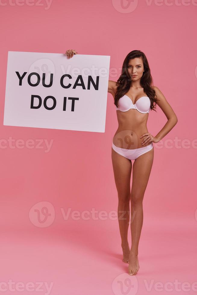 Follow your dreams.  Full length of attractive young woman holding a poster and keeping hand on hip while standing against pink background photo