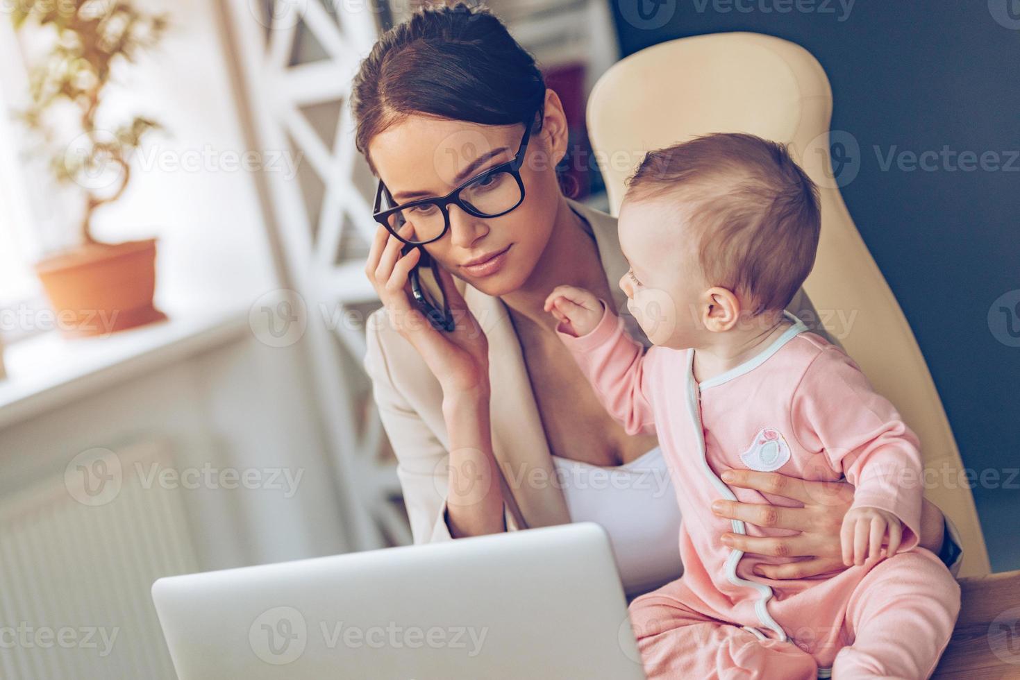 déjame hablar con tu socia de negocios, una joven y hermosa mujer de negocios hablando por teléfono móvil y mirando una laptop mientras está sentada con su bebé en su lugar de trabajo foto