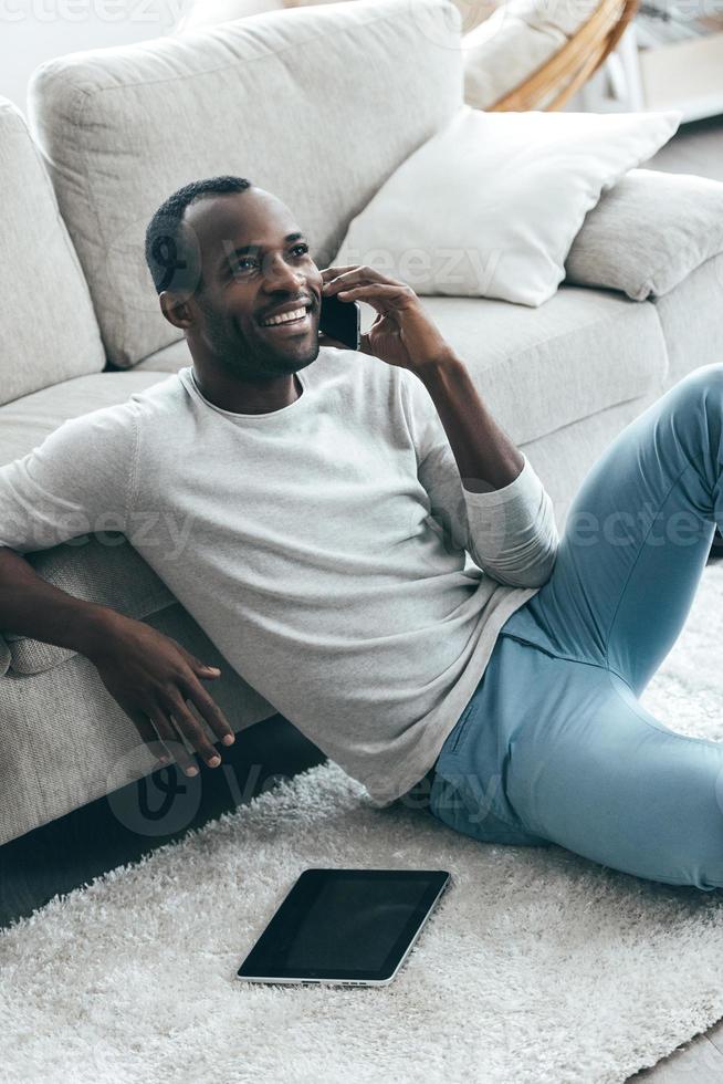 Great news Handsome young African man talking on the phone and smiling while sitting on the carpet at home photo
