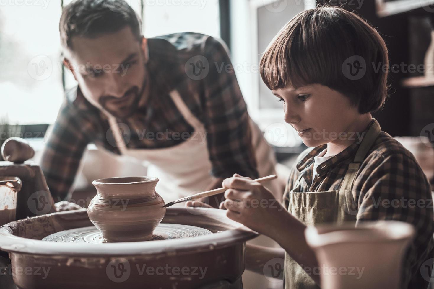 Creativity is in his veins. Little boy drawing on ceramic pot at the pottery class while man in apron standing close to him photo