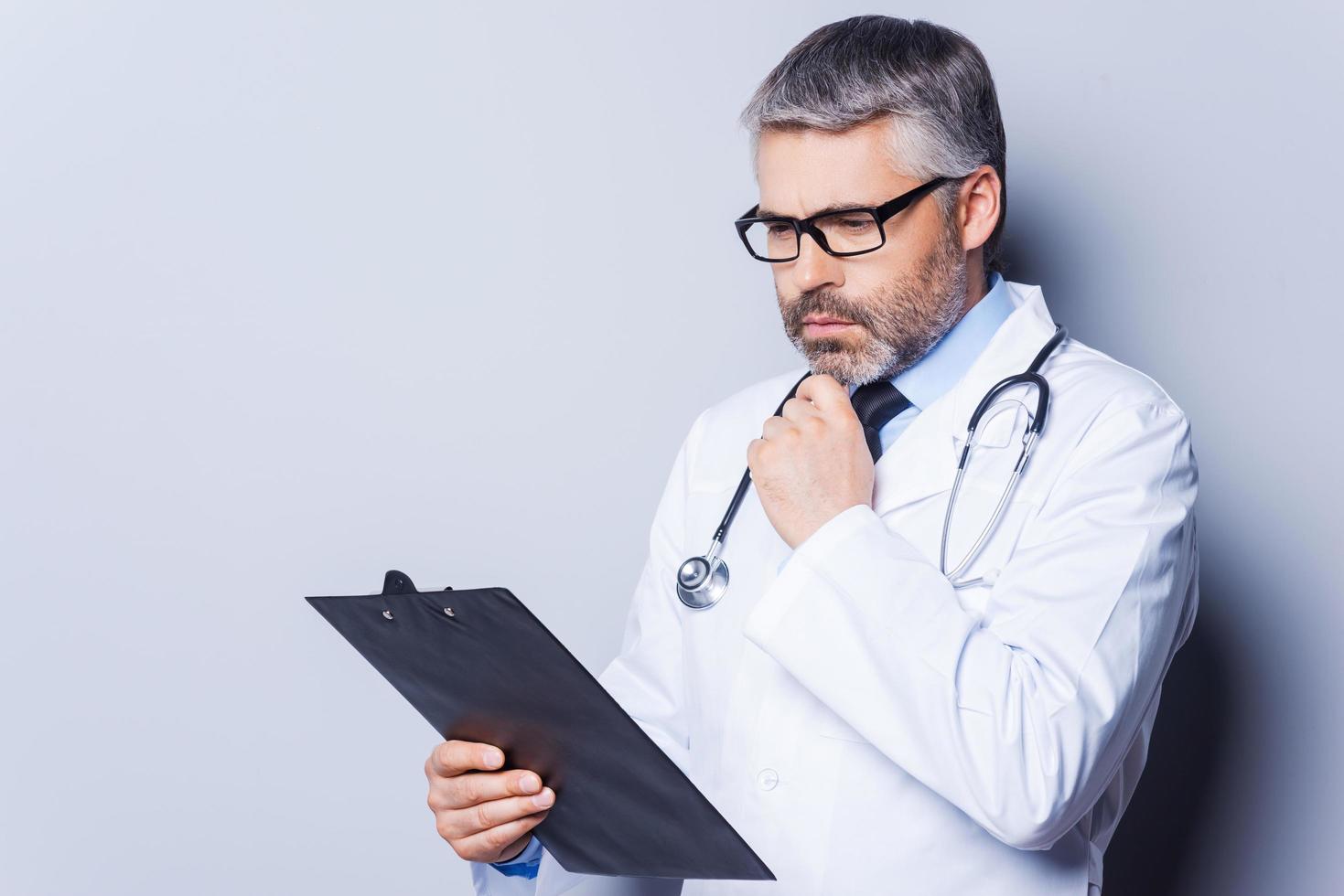 Thoughtful doctor. Thoughtful mature doctor holding hand on chin and looking at his clipboard while standing against grey background photo