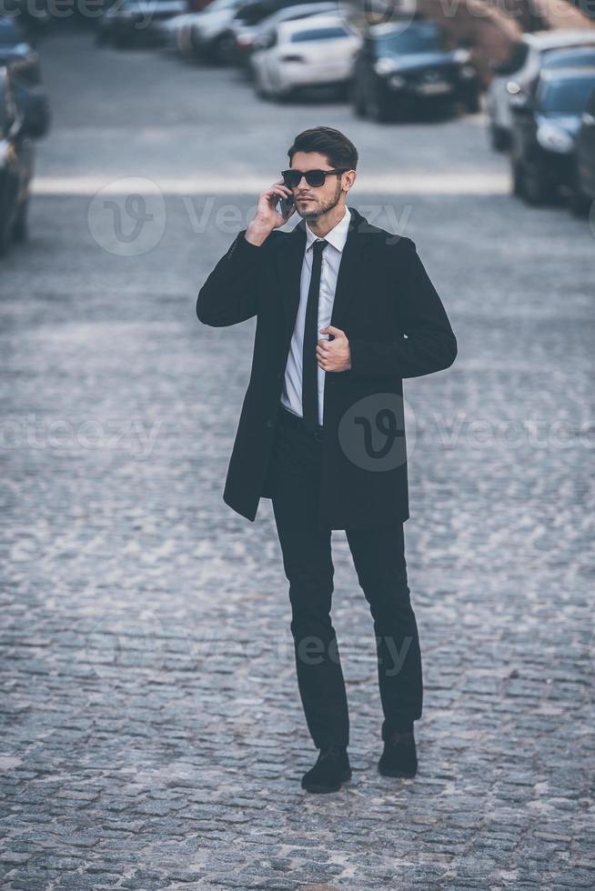 I will be in ten minutes Full length of handsome young well-dressed man in sunglasses talking on mobile phone and looking away while walking outdoors photo