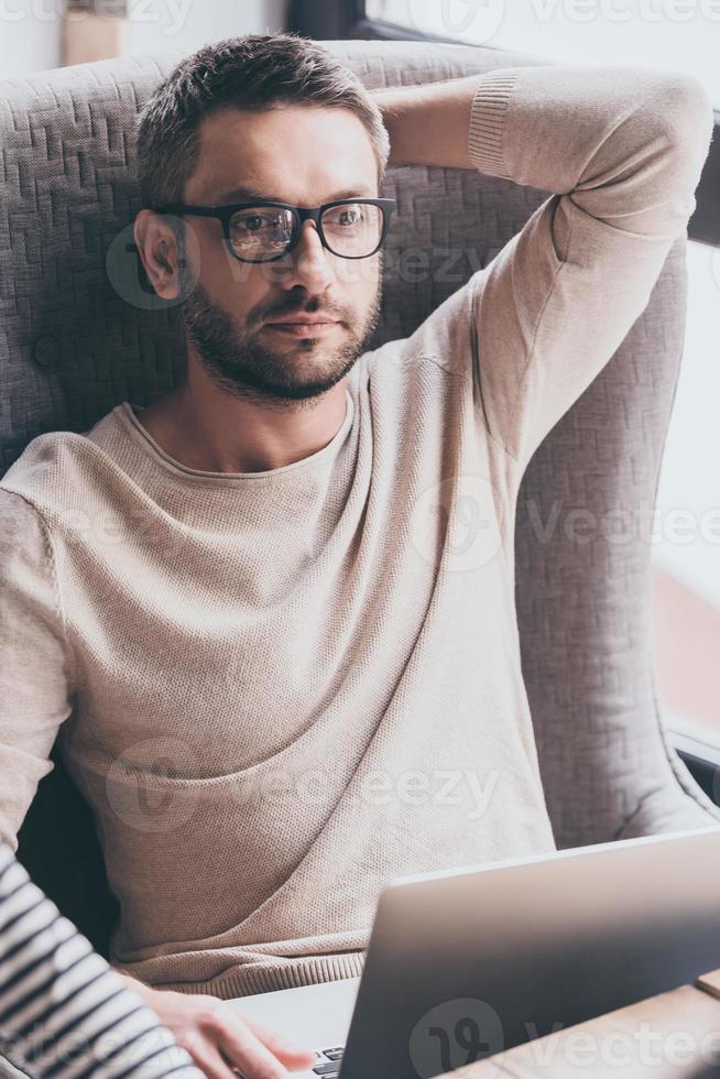 Handsome and thoughtful. Handsome man using his laptop and looking pensive while sitting in chair photo