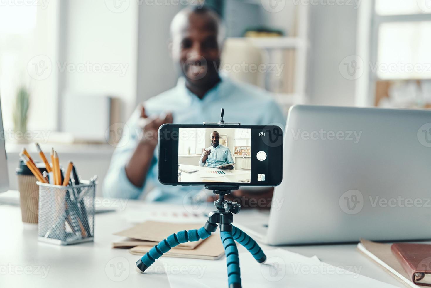 apuesto joven africano con camisa diciendo algo y sonriendo mientras hace un video en las redes sociales foto