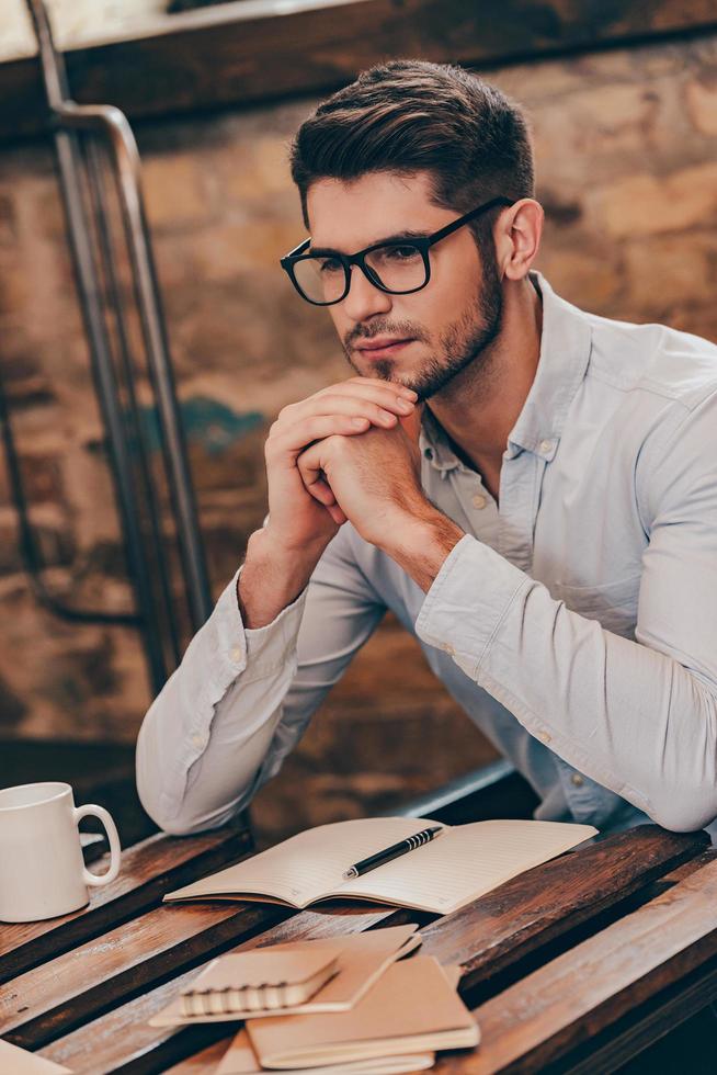 Thinking about solution. Handsome thoughtful young man keeping hands clasped and looking away while sitting at his working place photo