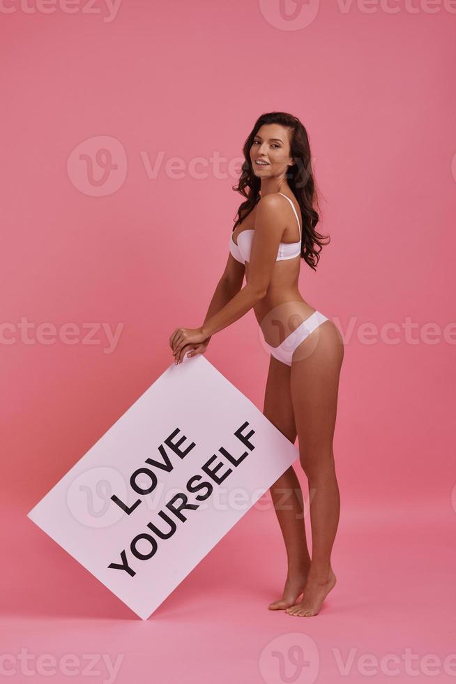Smile to yourself every day.  Full length of attractive young smiling woman holding a poster and looking at camera while standing against pink background photo